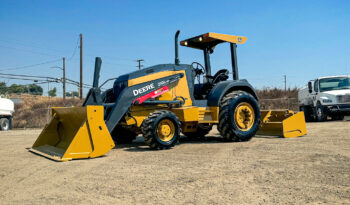 John Deere 210LEP Skip Loader parked on a dirt lot with construction vehicles in the background under a clear blue sky.