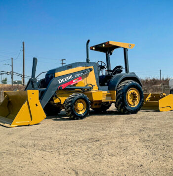 John Deere 210LEP Skip Loader parked on a dirt lot with construction vehicles in the background under a clear blue sky.