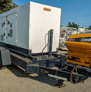 The image shows a towable Multiquip 180KVA generator mounted on a heavy-duty dual-axle trailer. The generator housing is white, with ventilation grilles and latches on the sides for access. The trailer has a black frame and it includes a hitch setup for towing, complete with a chain and heavy-duty hook. In the background, a yellow LeeBoy 8515 machine is visible, along with other industrial equipment. The scene appears to be in an outdoor yard or equipment lot.