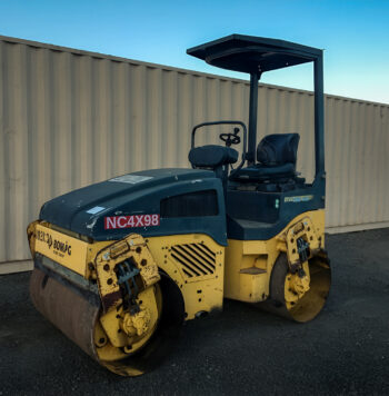 Bomag BW120AD-4 Vibratory Tandem Roller with Smooth Drum, featuring a compact design and an open canopy, parked on a dirt lot in front of a beige shipping container at sunset.