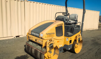 A Volvo DD25B Compactor with a smooth drum parked on a dirt lot in front of beige shipping containers under a clear blue sky.