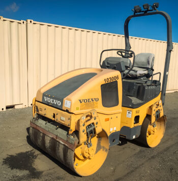 A Volvo DD25B Compactor with a smooth drum parked on a dirt lot in front of beige shipping containers under a clear blue sky.