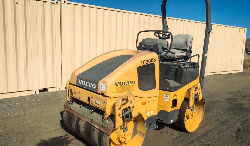 A Volvo DD25B Compactor with a smooth drum parked on a dirt lot in front of beige shipping containers under a clear blue sky.